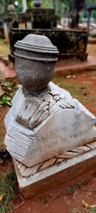 A gravestone in a cemetery features a sculpted urn on top. The stone is weathered with visible dirt and debris around the base. The inscription reads 'Slaap in vrede ontwaak in vreugde.' The surrounding environment includes grass and other tombstones.