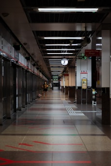 A long, empty train station hallway with a person walking and a large hanging clock displaying the time. The station features modern architecture with overhead lighting, signs, and a polished floor marked with red lines guiding passengers.