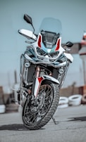 A modern, sleek motorcycle with a robust design is positioned upright on a paved surface. It features a large front wheel, dual headlights, and a windshield. The color scheme includes white and red accents with black details, and the reflective surface of the metal parts adds a shine.