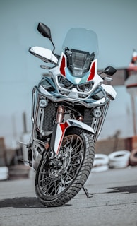 A modern, sleek motorcycle with a robust design is positioned upright on a paved surface. It features a large front wheel, dual headlights, and a windshield. The color scheme includes white and red accents with black details, and the reflective surface of the metal parts adds a shine.