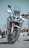 A modern, sleek motorcycle with a robust design is positioned upright on a paved surface. It features a large front wheel, dual headlights, and a windshield. The color scheme includes white and red accents with black details, and the reflective surface of the metal parts adds a shine.