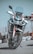 A modern, sleek motorcycle with a robust design is positioned upright on a paved surface. It features a large front wheel, dual headlights, and a windshield. The color scheme includes white and red accents with black details, and the reflective surface of the metal parts adds a shine.