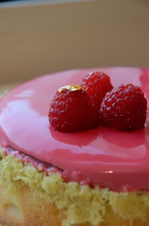 A round dessert topped with a smooth, shiny pink glaze and garnished with three fresh raspberries, one adorned with a small piece of gold leaf. The base of the dessert has a light, fluffy texture resembling sponge cake.