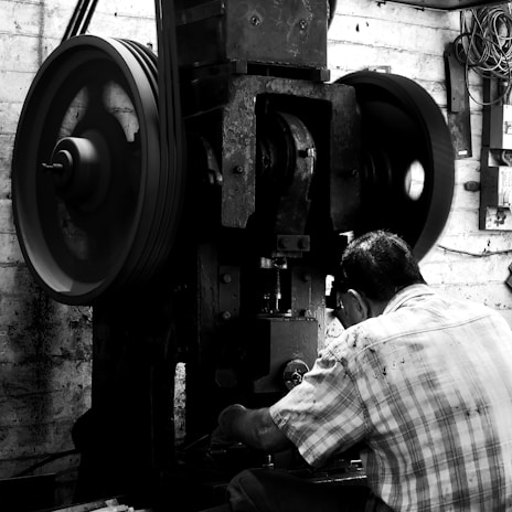 A person wearing a plaid shirt works on a large industrial machine with prominent wheels and belts. The setting appears to be inside a workshop with various tools and wires visible on the brick wall.