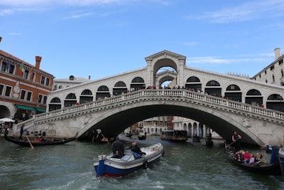 A large, ornate stone bridge spans a waterway, crowded with people walking across its arch. Below, several gondolas and motorboats navigate the canal. The surrounding architecture features classic Venetian style buildings with balconies.