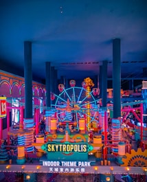 A vibrant and colorful indoor theme park with neon lights and a large Ferris wheel. The architecture features a playful, whimsical design with towering columns and various amusement rides. The signage prominently displays the name 'Skytropolis Indoor Theme Park' in both English and another language.
