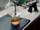 A glass filled with an amber-colored drink and ice sits on a black mat. A smoking gun and dome cover are used to infuse the drink with smoke. Various kitchen items, including a jar and mixing utensils, are seen on the marble countertop in the background.