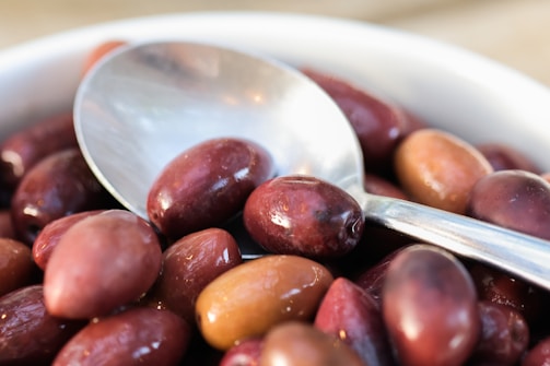 A close-up of a bowl filled with shiny, plump olives in varying shades of purple and brown. A metallic spoon rests atop the olives, reflecting some light.