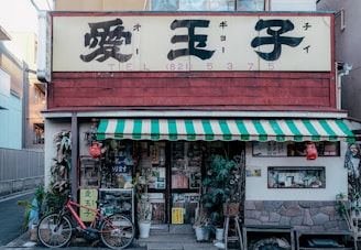 A small storefront with a traditional appearance, featuring Japanese signage above the entrance. The building has a red exterior with a green and white striped awning. A red bicycle is parked to the left, and various plants and decorations are visible in front of the shop. The windows display numerous posters and items, giving the space a cluttered aesthetic.