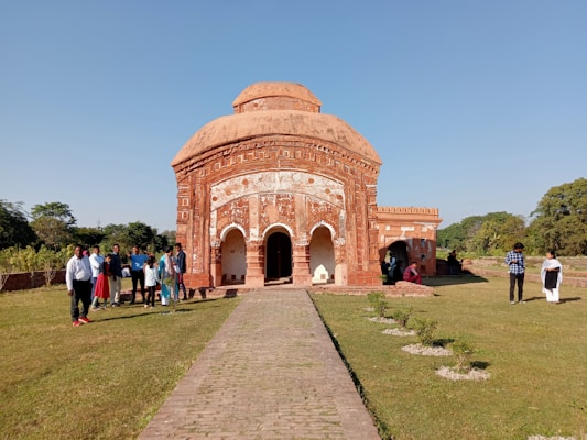 An ancient terracotta temple with intricate carvings stands under a clear blue sky. A group of people is gathered around the front, some dressed in traditional clothing, while others wear casual attire. The temple is set on a well-manicured lawn with a stone pathway leading to its entrance. Trees and greenery surround the area, creating a peaceful atmosphere.