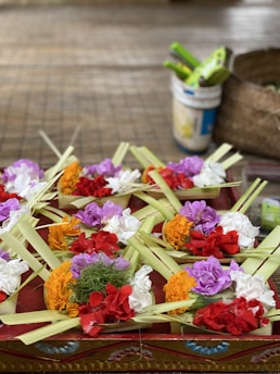 A collection of colorful flower offerings arranged with leaves on a decorative surface. Various flowers, including marigolds and carnations in red, orange, white, and purple hues, are neatly placed. In the background, there is a blurry view of a bucket and a woven basket.