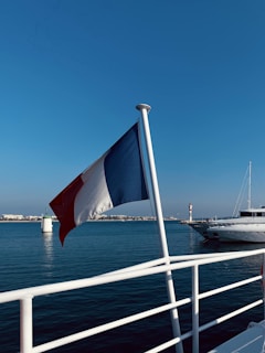 A French flag is prominently displayed on a boat with a calm sea backdrop. In the distance, a lighthouse and another white boat are visible against a clear blue sky.