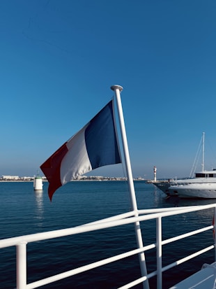 A French flag is prominently displayed on a boat with a calm sea backdrop. In the distance, a lighthouse and another white boat are visible against a clear blue sky.