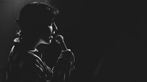 A person is singing into a microphone, captured in silhouette against a dark background. The lighting creates a dramatic effect, highlighting the contours of their face and the microphone.