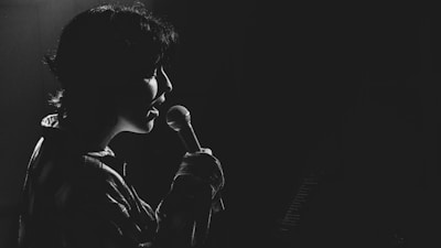 A person is singing into a microphone, captured in silhouette against a dark background. The lighting creates a dramatic effect, highlighting the contours of their face and the microphone.