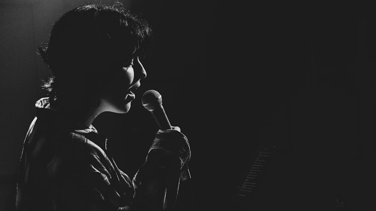 A person is singing into a microphone, captured in silhouette against a dark background. The lighting creates a dramatic effect, highlighting the contours of their face and the microphone.