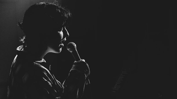 A person is singing into a microphone, captured in silhouette against a dark background. The lighting creates a dramatic effect, highlighting the contours of their face and the microphone.