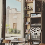 A cozy interior space featuring a vintage audio setup with large speakers, an amplifier, and reel-to-reel tape recorder on a wooden shelf. The room is adorned with small potted plants and various coffee-related objects, suggesting a café setting. Through large glass windows, an outdoor view of a stone building and green plants is visible, providing a historical and serene atmosphere.