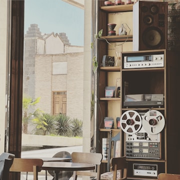 A cozy interior space featuring a vintage audio setup with large speakers, an amplifier, and reel-to-reel tape recorder on a wooden shelf. The room is adorned with small potted plants and various coffee-related objects, suggesting a café setting. Through large glass windows, an outdoor view of a stone building and green plants is visible, providing a historical and serene atmosphere.