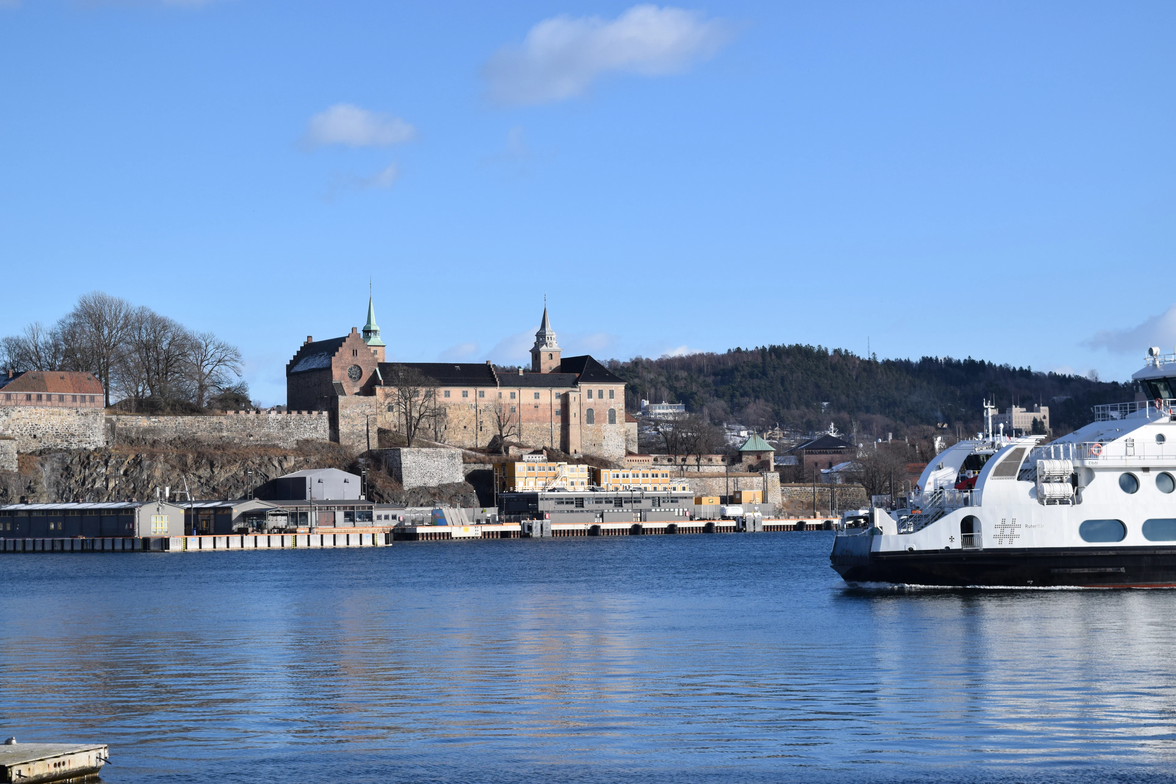 A white public fjord ferry crossing Oslo harbour in front of Akershus Fortress, the medieval ramparts and green-spired castle standing on their wooded bluff above the quays under a clear blue sky.