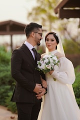 A couple is posing outdoors; the man is wearing a black suit and glasses, while the woman is dressed in a white wedding gown, holding a bouquet of white flowers. The background features greenery and a blurred structure, creating a soft, romantic setting.