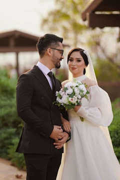 A couple is posing outdoors; the man is wearing a black suit and glasses, while the woman is dressed in a white wedding gown, holding a bouquet of white flowers. The background features greenery and a blurred structure, creating a soft, romantic setting.