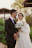A couple is posing outdoors; the man is wearing a black suit and glasses, while the woman is dressed in a white wedding gown, holding a bouquet of white flowers. The background features greenery and a blurred structure, creating a soft, romantic setting.
