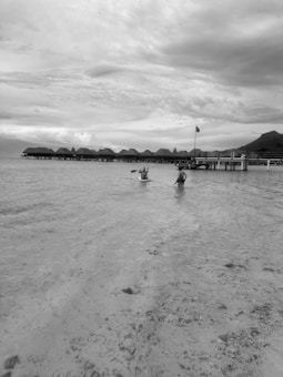 Overwater bungalows stretch across the horizon on a calm sea. Two kayakers paddle in the shallow, clear waters. The sky is filled with clouds, adding a dramatic atmosphere.