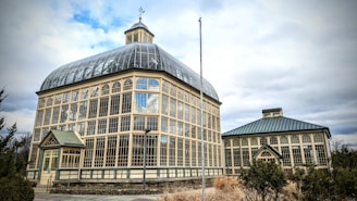 A large, historic greenhouse with a glass and metal framework stands prominently under a partly cloudy sky. The structure features numerous arched windows that allow light to stream into the interior. The main building has a rounded roof with a square tower on top, and there is a smaller, similar structured building adjacent to it. Surrounding the greenhouse are patches of shrubs and dry grass, hinting at a winter or early spring setting.