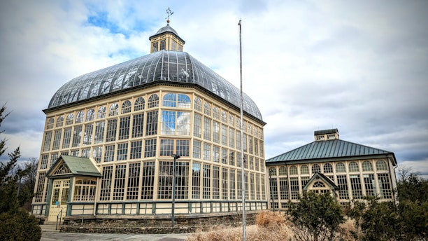 A large, historic greenhouse with a glass and metal framework stands prominently under a partly cloudy sky. The structure features numerous arched windows that allow light to stream into the interior. The main building has a rounded roof with a square tower on top, and there is a smaller, similar structured building adjacent to it. Surrounding the greenhouse are patches of shrubs and dry grass, hinting at a winter or early spring setting.
