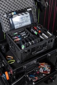 A black protective case is open, revealing an organized collection of electronic devices and accessories inside, such as labeled memory cards and a clapperboard. The case is situated on a rolling cart equipped with various colored carabiners and additional storage compartments.