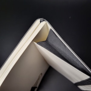 Close-up of the edge of an open notebook with ivory pages and a black elastic band. The pages are fanned slightly, revealing their texture and the precision of the binding. The notebook has a minimalist design, with the black band contrasting against the light pages.