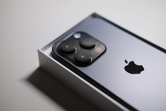 A close-up image of a sleek smartphone partially in its box, showcasing a metallic surface with three prominent camera lenses and an Apple logo.