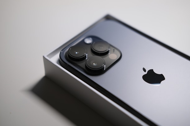 A close-up image of a sleek smartphone partially in its box, showcasing a metallic surface with three prominent camera lenses and an Apple logo.