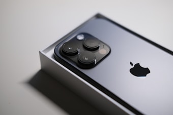 A close-up image of a sleek smartphone partially in its box, showcasing a metallic surface with three prominent camera lenses and an Apple logo.