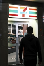 A person is walking towards the entrance of a well-lit convenience store with a recognizable logo and signage. The store interior is visible through the glass doors and windows, with shelves stocked with products.