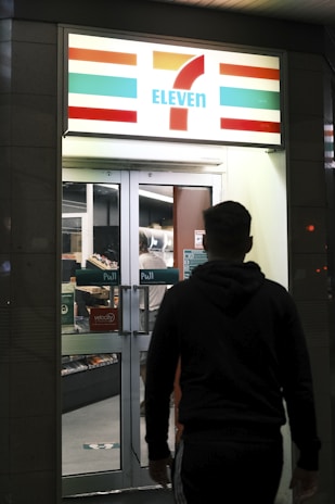 A person is walking towards the entrance of a well-lit convenience store with a recognizable logo and signage. The store interior is visible through the glass doors and windows, with shelves stocked with products.