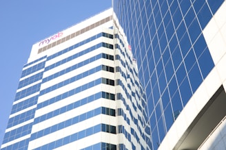 A modern skyscraper with reflective glass windows stands against a clear blue sky. The building features a series of horizontal stripes and a visible logo at the top.