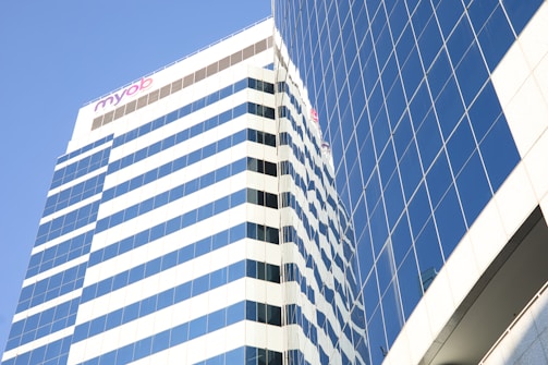 A modern skyscraper with reflective glass windows stands against a clear blue sky. The building features a series of horizontal stripes and a visible logo at the top.