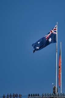 A blue sky serves as the backdrop for two large flags flying atop flagpoles. The flag on the left features a blue field with white stars and a Union Jack in the corner, commonly known as the flag of Australia. The flag on the right has a black and red field with a yellow circle in the center, known as the Aboriginal flag. Below the flags, several people in uniform line the top of a structure.