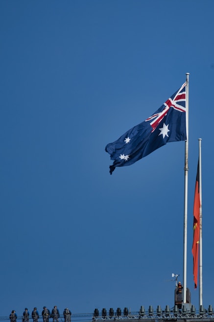 A blue sky serves as the backdrop for two large flags flying atop flagpoles. The flag on the left features a blue field with white stars and a Union Jack in the corner, commonly known as the flag of Australia. The flag on the right has a black and red field with a yellow circle in the center, known as the Aboriginal flag. Below the flags, several people in uniform line the top of a structure.