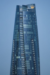 A modern skyscraper with a sleek, curved design and reflective glass facade that mirrors the clear blue sky. The building features a golden crown logo near the top.