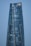 A modern skyscraper with a sleek, curved design and reflective glass facade that mirrors the clear blue sky. The building features a golden crown logo near the top.