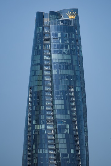 A modern skyscraper with a sleek, curved design and reflective glass facade that mirrors the clear blue sky. The building features a golden crown logo near the top.