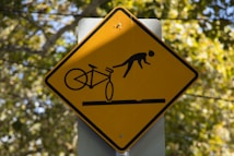 A yellow, diamond-shaped warning sign indicates a hazard for cyclists. It features a stylized image of a cyclist and a bicycle tipping forward, suggesting a potential accident or obstacle.