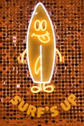 A neon light in the shape of a smiling surfboard character with arms and legs, on a background made of shiny, circular sequins. The words 'Surf's Up' are displayed beneath the surfboard.