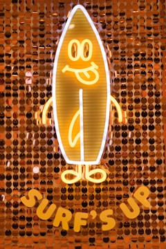 A neon light in the shape of a smiling surfboard character with arms and legs, on a background made of shiny, circular sequins. The words 'Surf's Up' are displayed beneath the surfboard.
