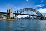 A large arched steel bridge stretches across a wide body of water, connecting two urban areas. The skyline features modern high-rise buildings partially obscured by the bridge. In the foreground, several boats are visible on the water's surface, which reflects the structures and clear blue sky above. A smaller sandstone-colored building and lush greenery are located near one end of the bridge.