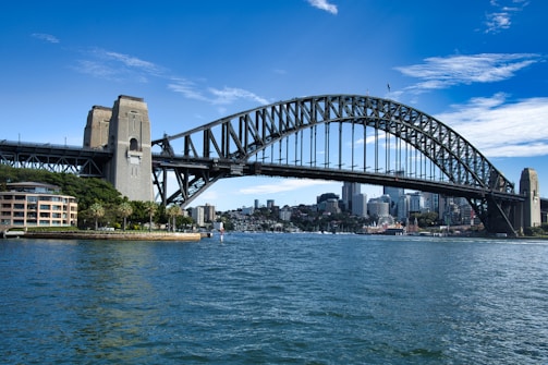 A large arched steel bridge stretches across a wide body of water, connecting two urban areas. The skyline features modern high-rise buildings partially obscured by the bridge. In the foreground, several boats are visible on the water's surface, which reflects the structures and clear blue sky above. A smaller sandstone-colored building and lush greenery are located near one end of the bridge.