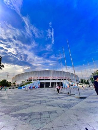 A modern architectural structure with the words 'Jakarta International Velodrome' displayed prominently. The building is surrounded by several tall flagpoles, and the foreground features a large tiled plaza. The sky is vibrant with blue and white clouds, creating a dramatic backdrop. Several people are scattered across the area, suggesting a public or leisurely setting.